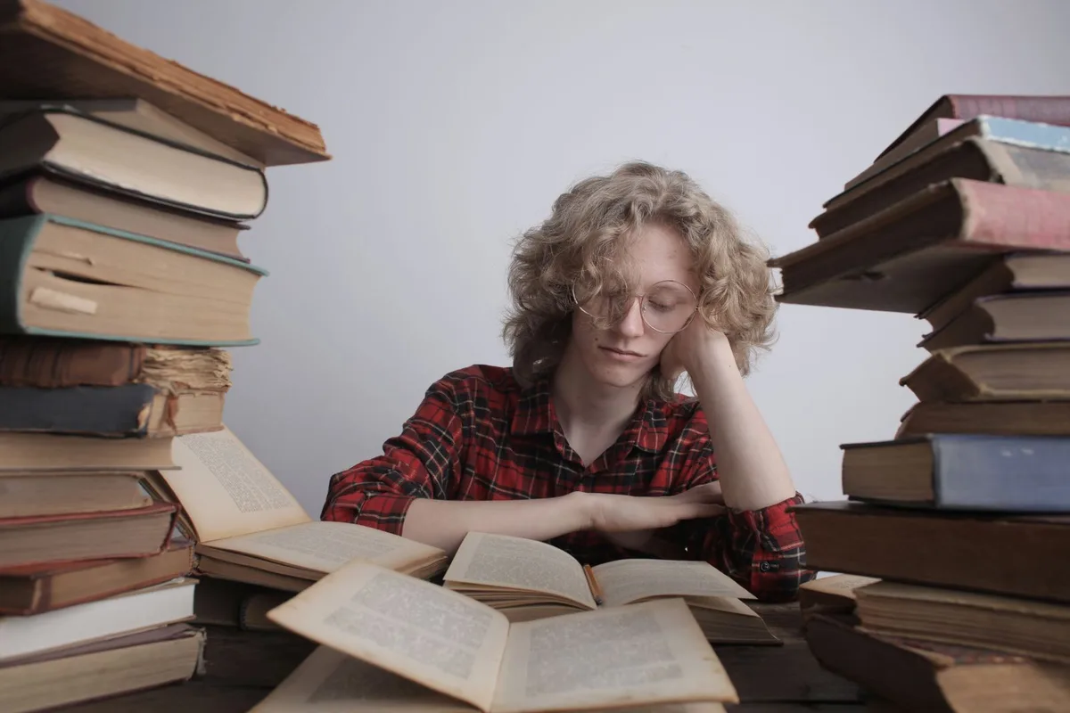 Уставший подросток сидит среди стопок раскрытых книг