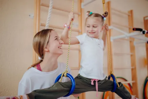 Улыбающаяся девочка висит на гимнастических кольцах в зале со шведской стенкой, рядом сидит молодая женщина